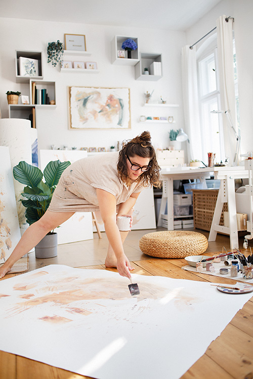  Rose Reichart malend in ihrem hellen lichtdurchflutenden Atelier. Die Künstlerin ist in dynamisch tänzerischer Bewegung getroffen und bringt gerade ihre handgemachte Farbe auf dem großformatigen Kunstwerk auf. Pinsel, Palette, Farbgläser, Bunstifte, Skizzenbücher sowie angefangene Kunstwerke unterstreichen die lebendige Atmosphäre des Kunst-Studios. Diverse Pflanzen sorgen für weitere farbige Akzente am inspirierenden Kreativ Arbeitsplatz.