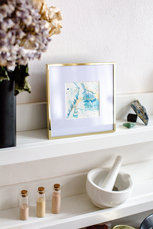  Ein kleines gerahmtes Kunstwerk von Rose Reichart auf einem gestylten Regal mit maritimem Boho Flair. Blau, Gold und Weiß prägen das abstrakte Gemälde. Kristalle, Pigmentfläschchen, getrocknete Blumen und ein Mörser mit Stößel sind auf dem weißen, schwebenden Holz ausgestellt.