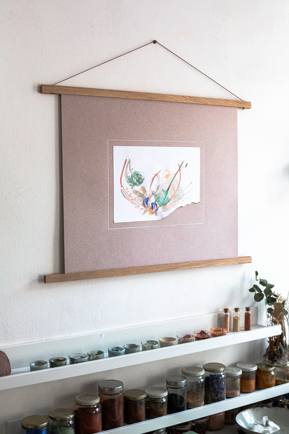 The light-filled art studio of artist Rose Reichart, featuring one of her paintings, compassion i. On the shelf below are her handcrafted paints which she mixed using natural earth pigments. The colorful glasses are organized on white picture rails. An aesthetic art studio organization hack for your small items that make photographing your art easy and professional.