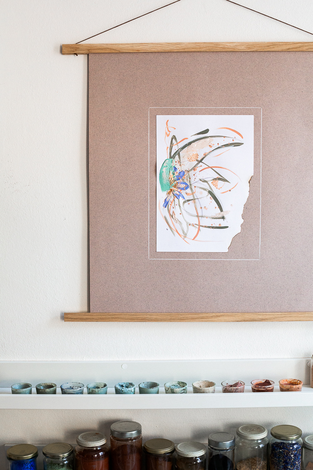 The light-filled art studio of artist Rose Reichart, featuring one of her paintings, compassion iii. On the shelf below are her handcrafted paints which she mixed using natural earth pigments. The colorful glasses are organized on white picture rails. An aesthetic art studio organization hack for your small items that make photographing your art easy and professional.