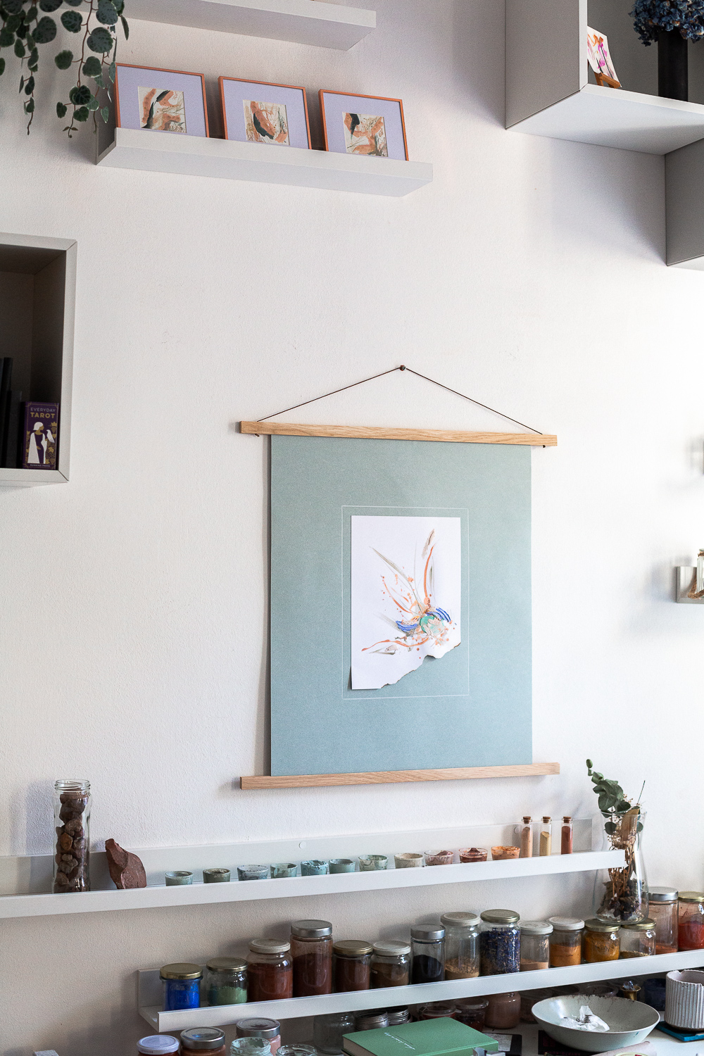 The light-filled art studio of artist Rose Reichart, featuring one of her paintings, perseverance i. On the shelf below are her handcrafted paints which she mixed using natural earth pigments. The colorful glasses are organized on white picture rails. An aesthetic art studio organization hack for your small items that make photographing your art easy and professional.