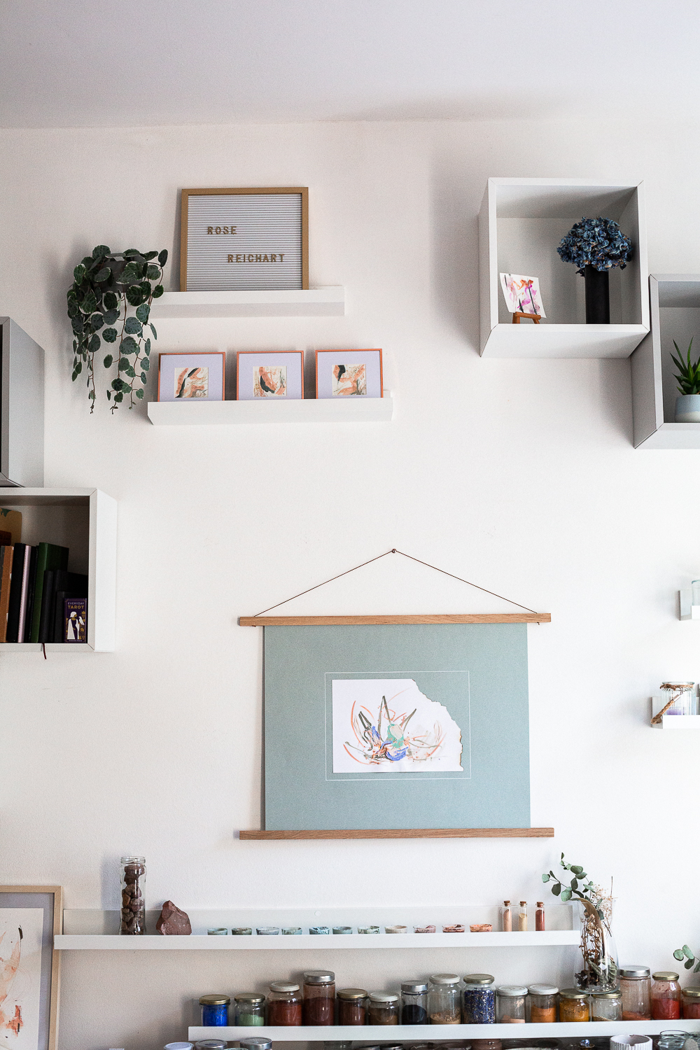The light-filled art studio of artist Rose Reichart, featuring one of her paintings, perseverance ii. On the shelf below are her handcrafted paints which she mixed using natural earth pigments. The colorful glasses are organized on white picture rails. An aesthetic art studio organization hack for your small items that make photographing your art easy and professional.