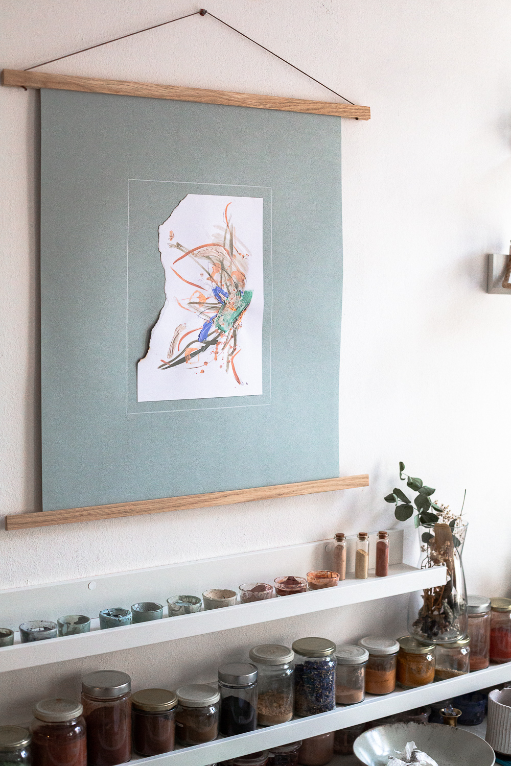 The light-filled art studio of artist Rose Reichart, featuring one of her paintings, perseverance iii. On the shelf below are her handcrafted paints which she mixed using natural earth pigments. The colorful glasses are organized on white picture rails. An aesthetic art studio organization hack for your small items that make photographing your art easy and professional.