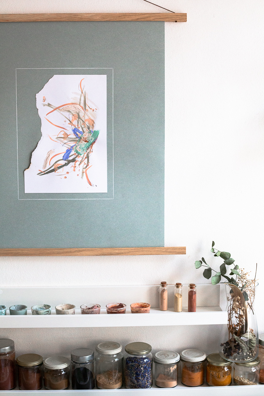 The light-filled art studio of artist Rose Reichart, featuring one of her paintings, perseverance iii. On the shelf below are her handcrafted paints which she mixed using natural earth pigments. The colorful glasses are organized on white picture rails. An aesthetic art studio organization hack for your small items that make photographing your art easy and professional.