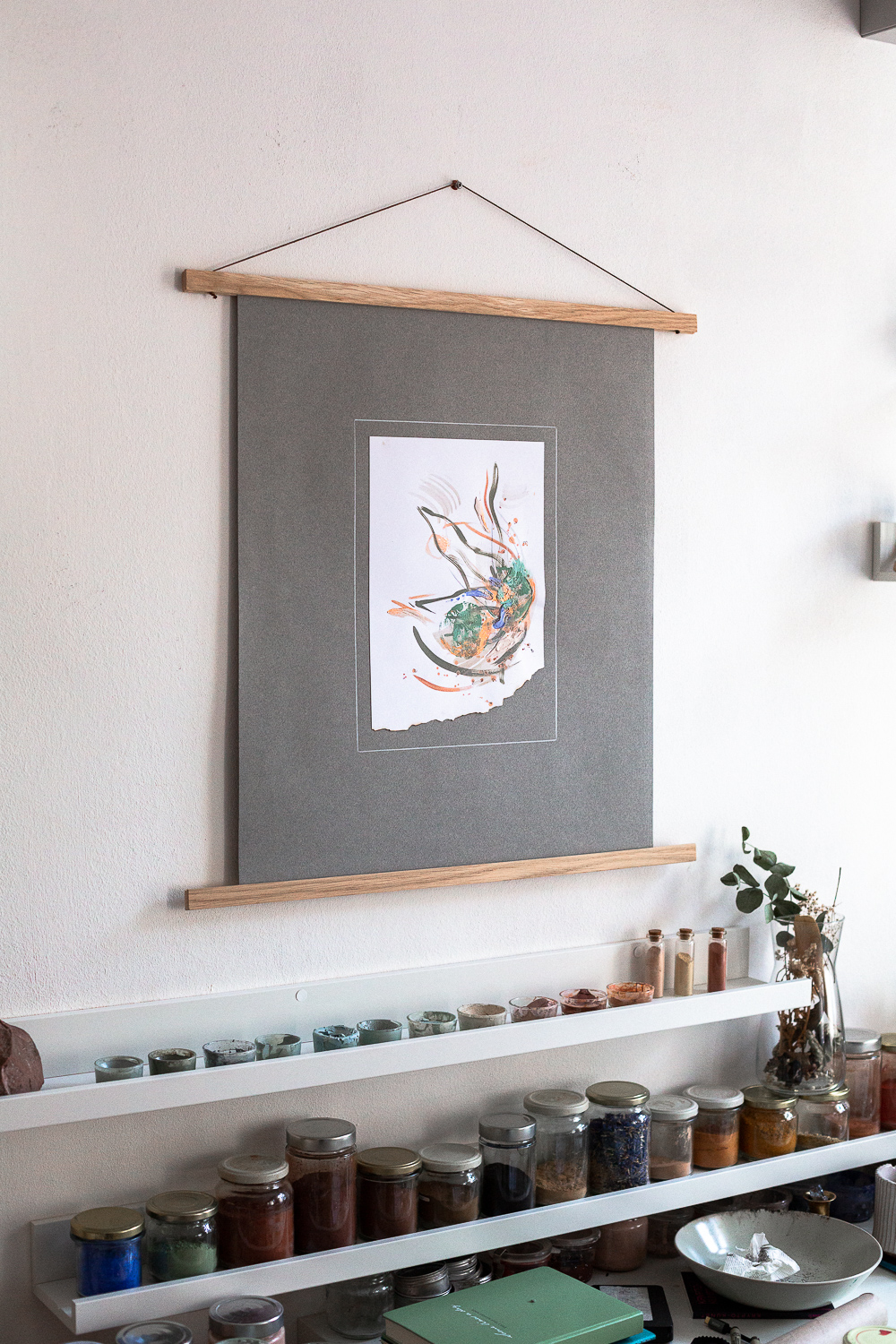 The light-filled art studio of artist Rose Reichart, featuring one of her paintings, solidarity iii. On the shelf below are her handcrafted paints which she mixed using natural earth pigments. The colorful glasses are organized on white picture rails. An aesthetic art studio organization hack for your small items that make photographing your art easy and professional.
