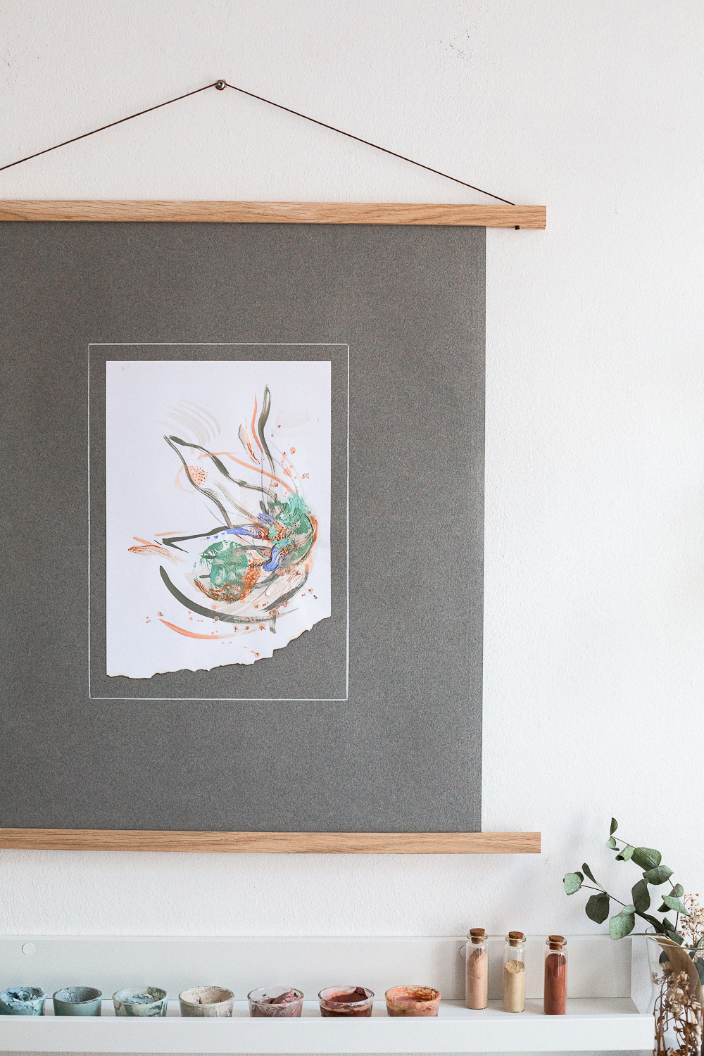 The light-filled art studio of artist Rose Reichart, featuring one of her paintings, solidarity iii. On the shelf below are her handcrafted paints which she mixed using natural earth pigments. The colorful glasses are organized on white picture rails. An aesthetic art studio organization hack for your small items that make photographing your art easy and professional.